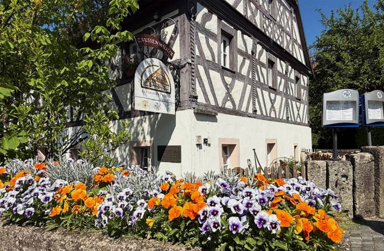 Fachwerkfassade der Zaussenmühle mit Wirtshausschild, davor ein Beet mit Stiefmütterchen