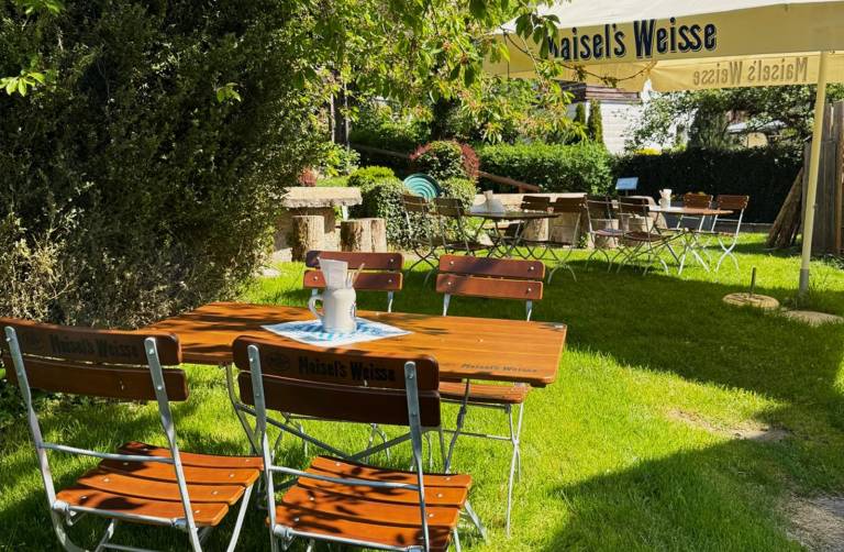 Holztische und Klappstühle unter einem Sonnenschirm auf der Wiese im Biergarten der Zaussenmühle