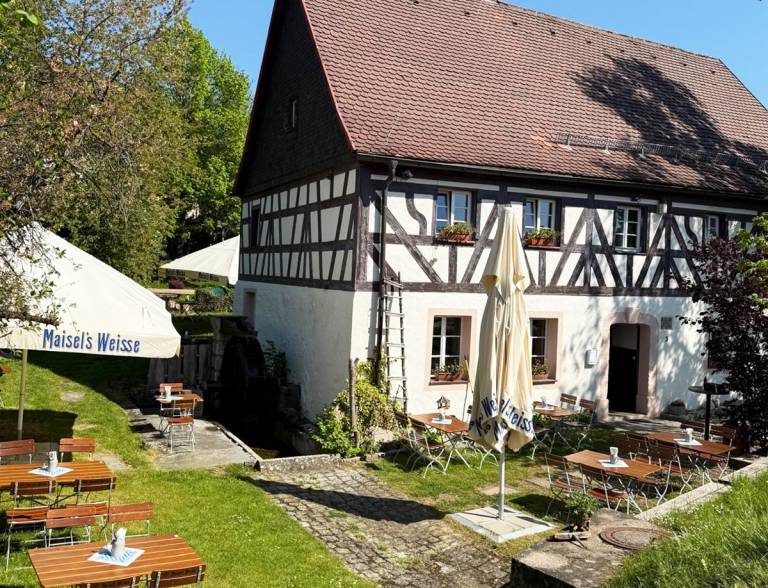 Biergarten mit Holztischen und Sonnenschirmen vor der Fachwerkfassade der Zaussenmühle in Pegnitz