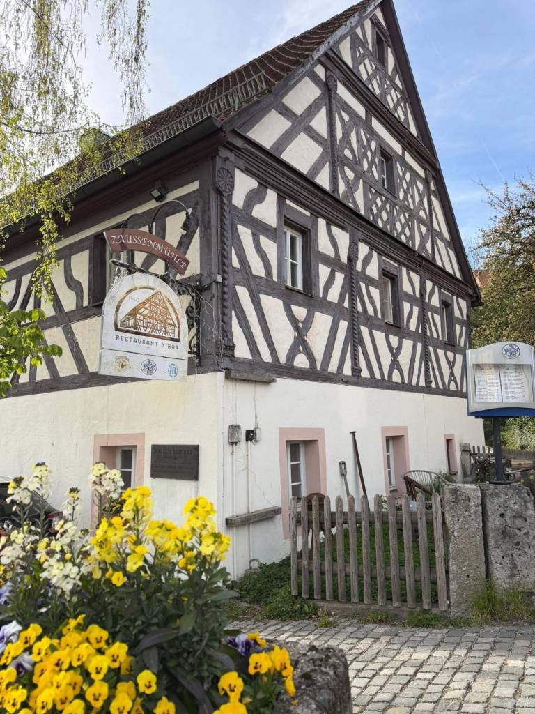 Eingangsseite der Zaussenmühle mit Fachwerkgiebel, Wirtshausschild und gelben Blumen im Vordergrund