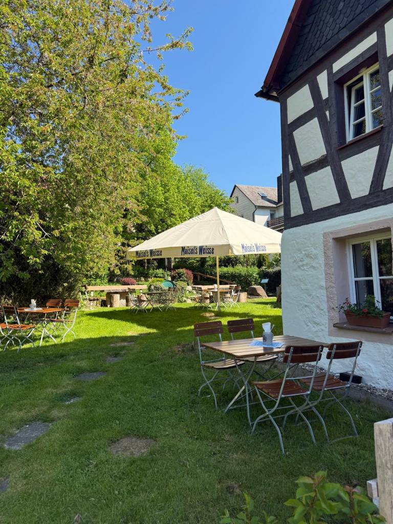 Tische und Stühle unter Sonnenschirmen im sommerlichen Biergarten der Zaussenmühle, daneben die Fachwerkfassade